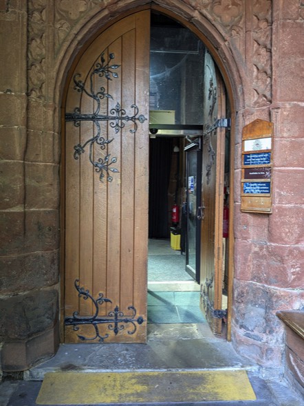A photograph showing a yellow ramp to the step into St Mary’s.  Large double wooden doors, the right-hand door is open.  Inside we can see one of a pair of automatic glass doors