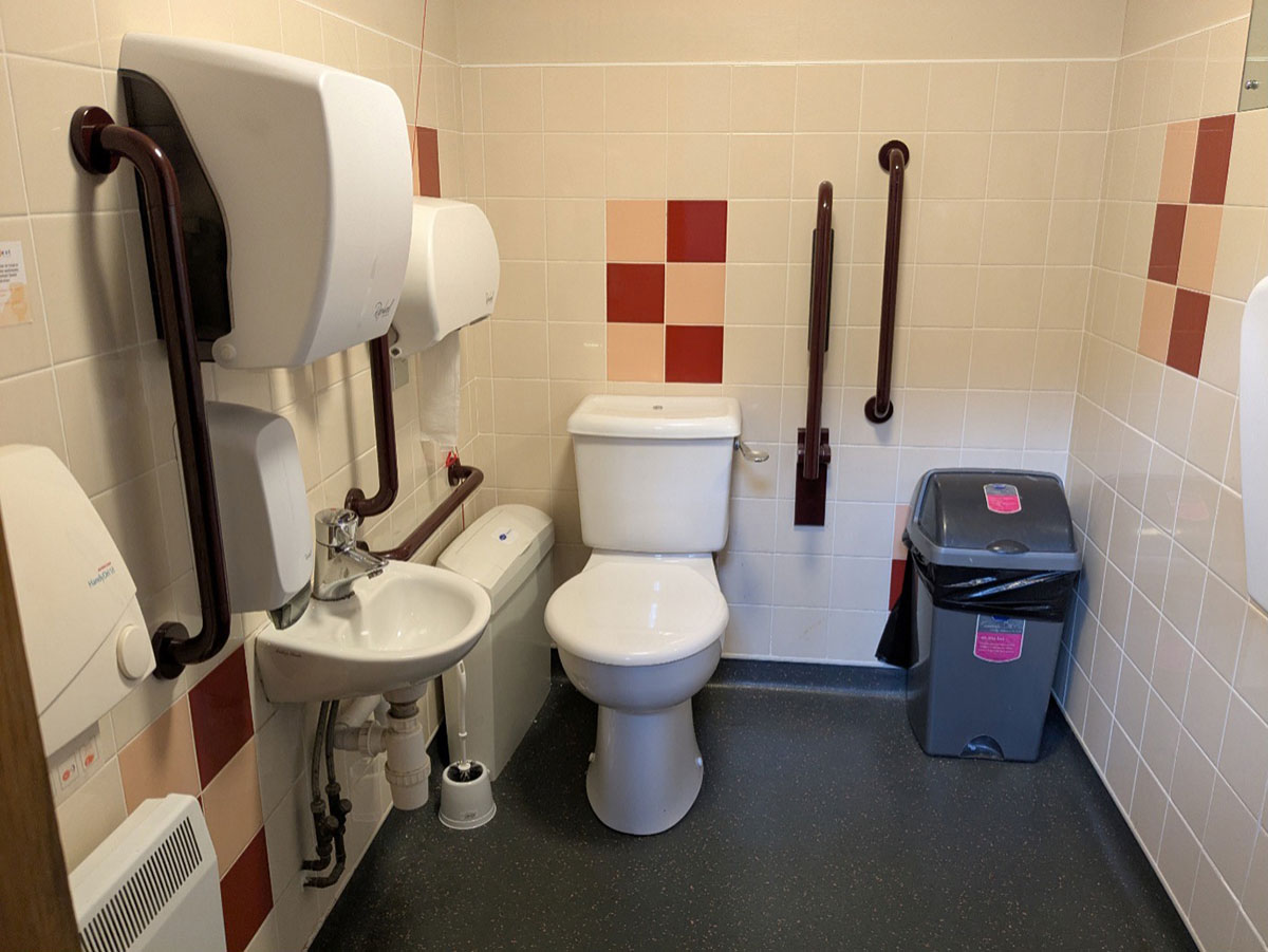 A photograph taken from the doorway, showing the interior of the accessible toilet.  The space is tiled with a vinyl floor.  Along the left wall, from the doorway, is a radiator with a hand-dryer above, a small basin with paper towel dispenser and soap dispenser above.  There is a vertical, wall mounted grab rail on each side of the basin.  Then a toilet brush and sanitary bin with a horizontal grab rail above it and above that a toilet roll dispenser and an emergency pull cord.  On the back wall from left to right is a toilet, a drop-down rail, a vertical, wall mounted grab rail and a grey flip top bin.  Along the right-hand wall, from the back of the room, is a wall mounted, fold up baby change station